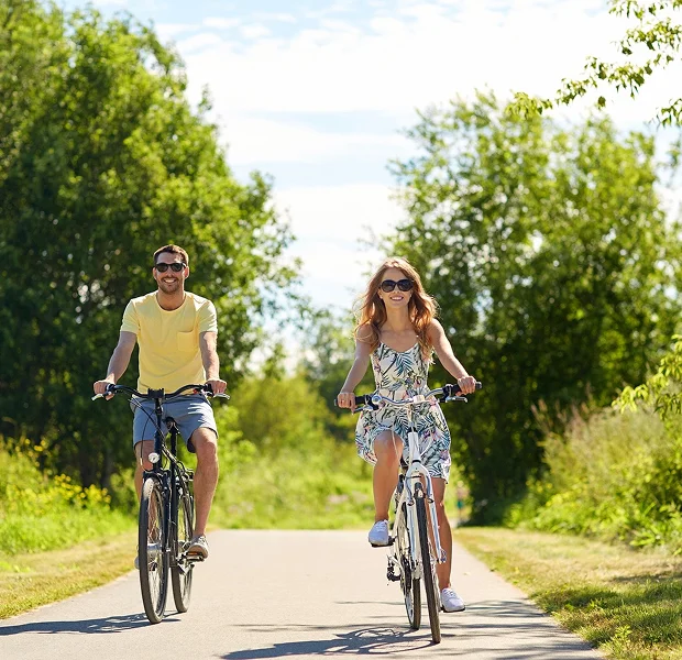 Couple souriant à vélo sur un chemin ensoleillé, entouré d'arbres.